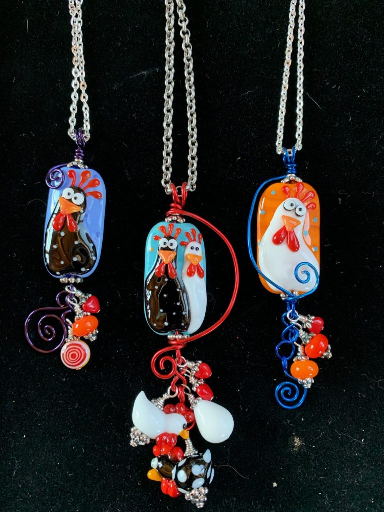 Glass beads by featuring cartoonish-looking portraits of rabbits, greyhounds, English bulldog, and chickens made with glass with colorful glass dangles, hanging from a chain by Melanie Schow